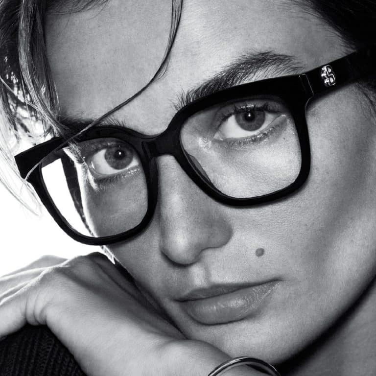 תקריב בשחור-לבן של אדם עם משקפיים גדולים, מביט בכוונה, ידו מונחת ליד פניו, נקודת חן בולטת.