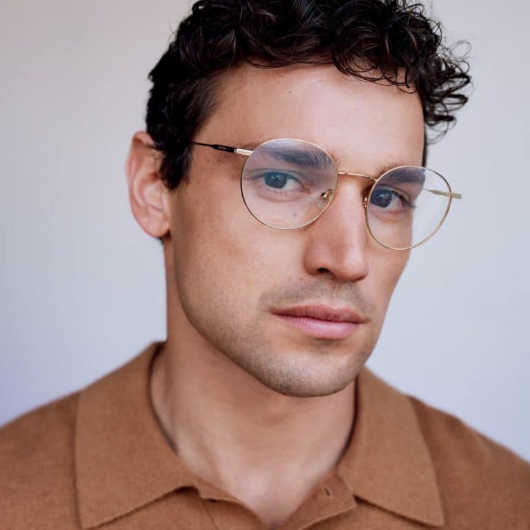 גבר עם שיער קצר ומתולתל ומשקפיים עגולים לובש חולצה חומה עם צווארון, ומביט מעט לצד.